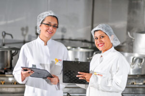 nutricionistas de jaleco, touca e prancheta sorriem em cozinha industrial da empresa Master Kitchen.