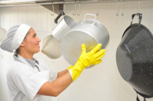 trabalhadora sorridente com luvas de limpeza e touca organizando panelas de aço inoxidavel na cozinha industrial