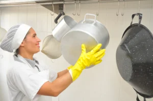 trabalhadora sorridente com luvas de limpeza e touca organizando panelas de aço inoxidavel na cozinha industrial